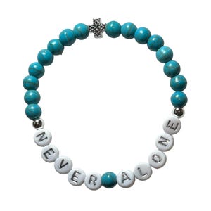 Könnte beinhalten: Ein türkisfarbenes Perlenarmband mit den Worten "NEVER ALONE" in weißen Buchstabenperlen. Ein kleines silbernes Kreuz und silberne Perlen akzentuieren das Design. Das Armband befindet sich auf weißem Hintergrund.