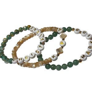 Peut inclure: Trois bracelets de perles. Un bracelet présente des perles vertes et jaunes, un autre des perles beiges et le troisième des perles vertes. Les bracelets ont des perles de lettres blanches épelant "CHRIST", "LOVE" et "KARMA".