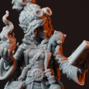 May include: A gray resin miniature figure of a character wearing goggles on their head, a long coat with a belt featuring a wheel emblem, and pouches. The figure holds a flame in one hand and an open book in the other, with a small creature on one shoulder.