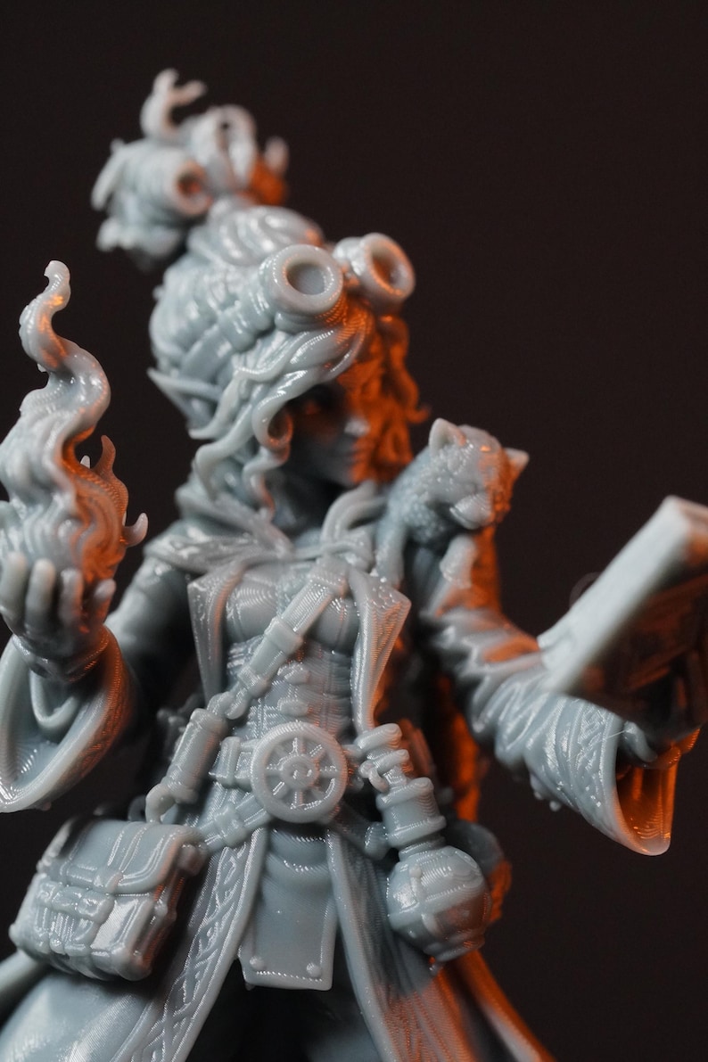 May include: A gray resin miniature figure of a character wearing goggles on their head, a long coat with a belt featuring a wheel emblem, and pouches. The figure holds a flame in one hand and an open book in the other, with a small creature on one shoulder.