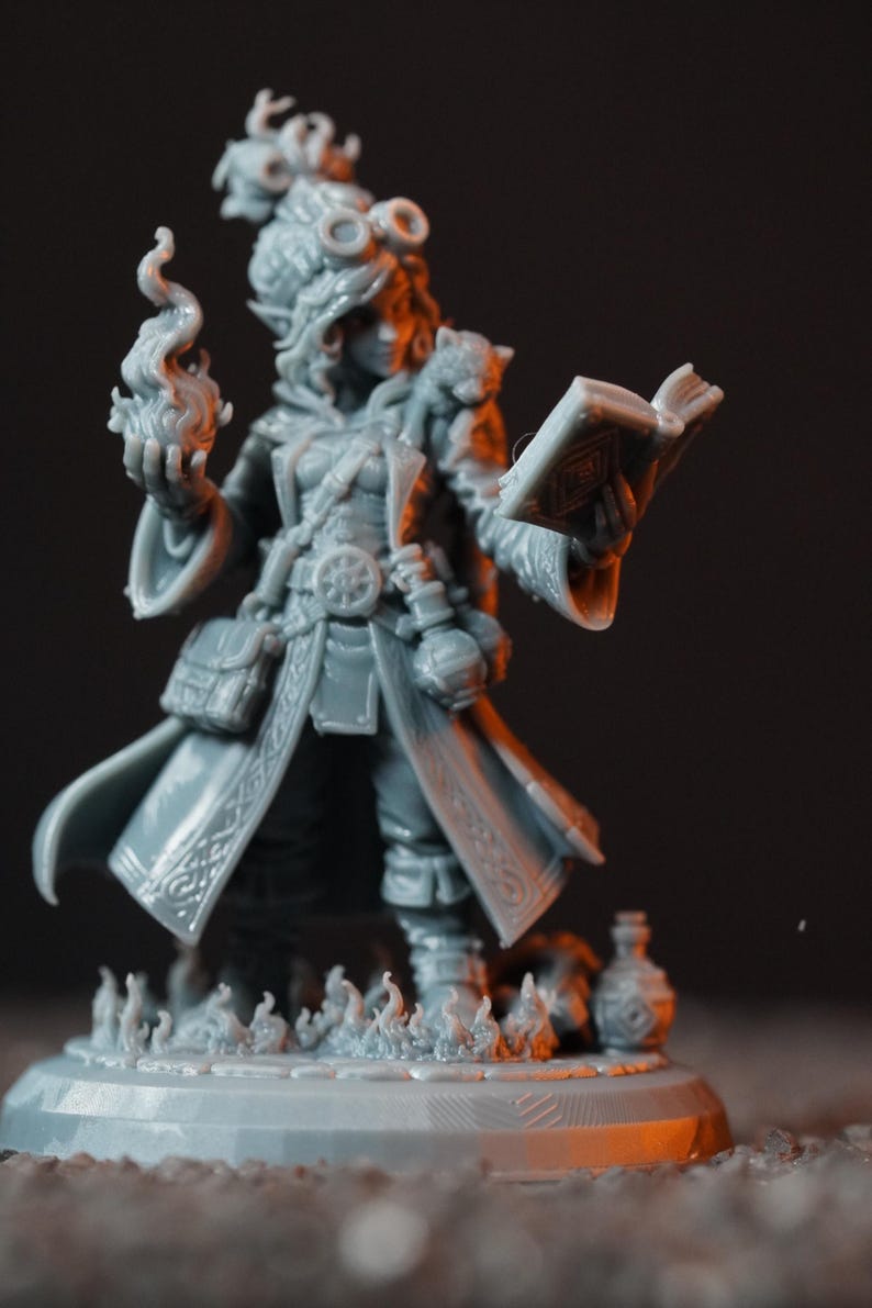May include: A gray unpainted resin miniature of a spellcaster figure wearing a long coat, goggles, and a belt with pouches. The figure holds a sculpted flame in one hand and an open book in the other, standing on a circular base with flame details.