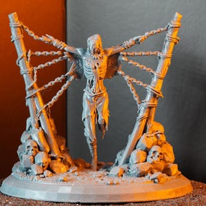 Op de afbeelding: Een grijs, gedetailleerd beeldje van een skeletfiguur gebonden aan een houten structuur met kettingen. De figuur heeft uitgestrekte armen en verscheurde kleding. Schedels en rotsen bevinden zich aan de basis, wat een gotisch of fantasiethema creëert.