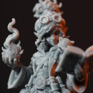 May include: A gray resin miniature figurine of a character wearing goggles on their head, holding an open book in one hand and a sculpted flame in the other. The figure features detailed clothing with a belt buckle and various small pouches.
