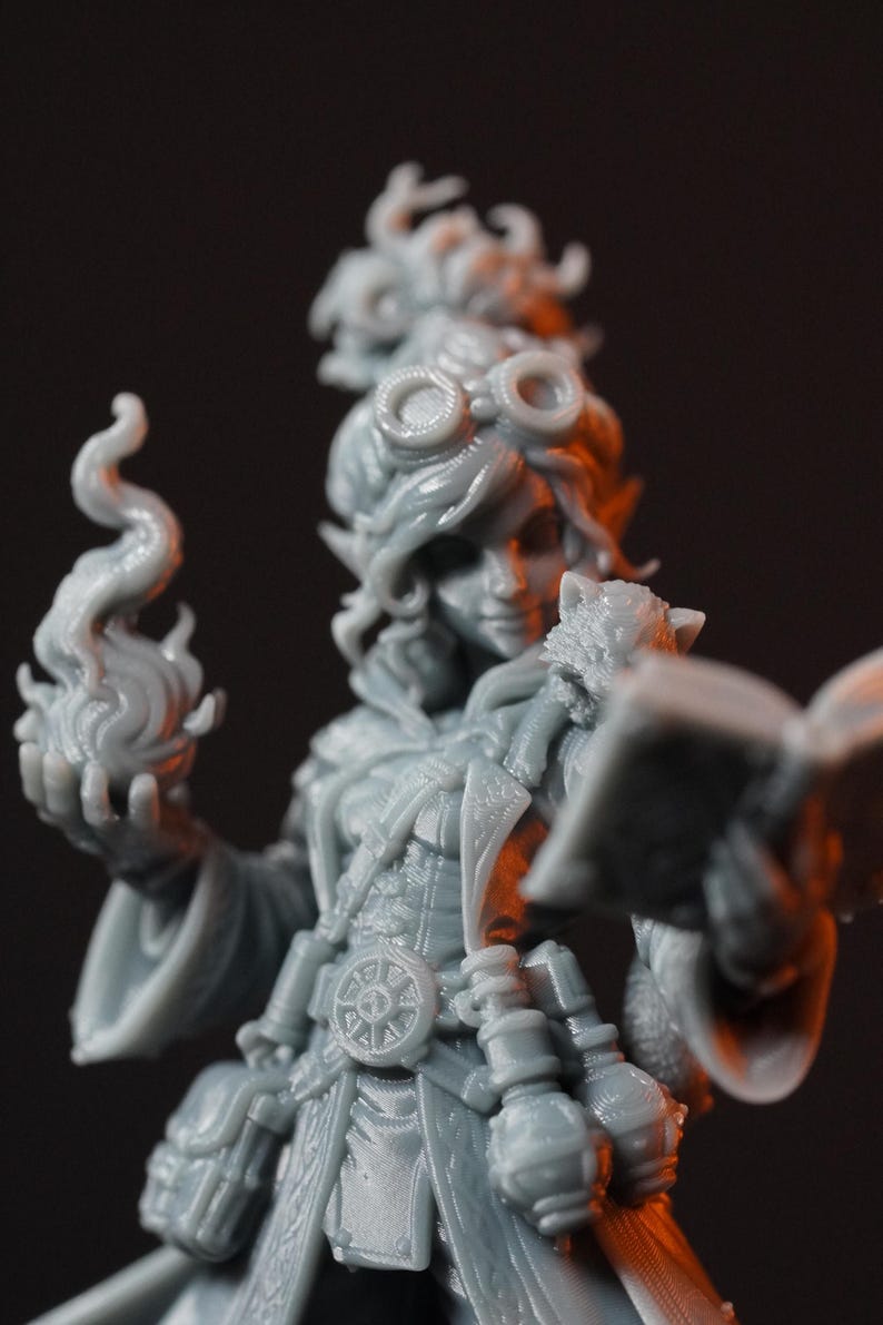 May include: A gray resin miniature figurine of a character wearing goggles on their head, holding an open book in one hand and a sculpted flame in the other. The figure features detailed clothing with a belt buckle and various small pouches.