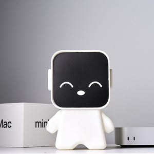 May include: A white, smiling robot-shaped speaker with a black screen face and small arms and legs. A white box with "Mac mini" printed on it and a silver device are also in the image.