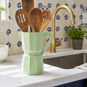 May include: A light green, geometric-shaped utensil holder in the style of a stovetop espresso maker, filled with wooden spoons and spatulas. The kitchen setting features a gold faucet, a marble countertop, and blue and white tile backsplash.