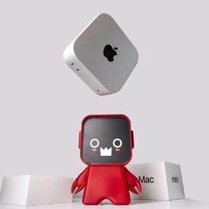 May include: A silver square computer with a black Apple logo floating above a red character figurine with a black screen face featuring white eyes and a crown-shaped mouth. Two white boxes labeled Mac mini are positioned behind the red figurine.