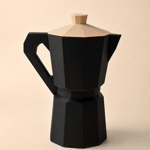 May include: A matte black moka pot with a faceted, geometric body and a handle. The lid is a light beige, faceted, and geometric shape with a small knob on top. The overall design features sharp, angular lines and a distinct octagonal structure.