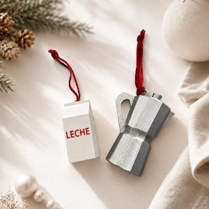 May include: Two hanging ornaments featuring a white milk carton with the red text LECHE and a silver-toned metallic moka pot coffee maker. Each ornament is attached to a thin red velvet ribbon for hanging, designed as decorative holiday accents.