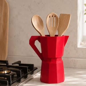May include: A bright red, faceted utensil holder shaped like a moka pot, containing three light-colored wooden kitchen tools: a solid spoon, a slotted spoon, and a flat spatula. The container features a geometric design with a handle and spout.