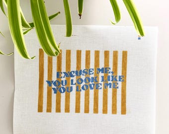 May include: A white needlepoint canvas featuring vertical mustard yellow stripes and the blue embroidered text "EXCUSE ME, YOU LOOK LIKE YOU LOVE ME" centered across the striped pattern. The canvas has a grid-like texture throughout.