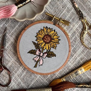 May include: An embroidered sunflower design in a wooden hoop. The sunflower is yellow and brown with green leaves and a pink bow. Embroidery floss, needles, and scissors are arranged around the hoop, suggesting a craft project.