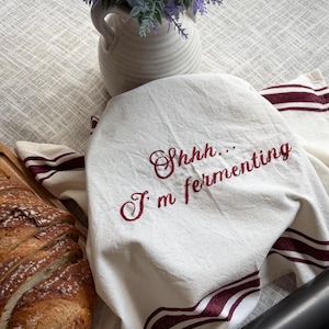 May include: A white kitchen towel with burgundy stripes and embroidered text reading "Shhh... I'm fermenting" covers a bowl. A loaf of sliced bread rests on a wooden board, and a vase of purple flowers sits in the background.