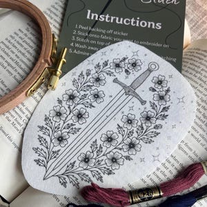 May include: A white fabric patch with a black floral and sword design, surrounded by embroidery floss and a wooden hoop. The patch has the text "Stick and Stitch Instructions".