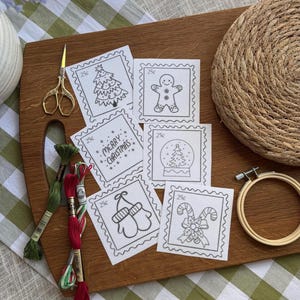 May include: A wooden cutting board displays Christmas-themed embroidery patterns, including a Christmas tree, gingerbread man, and candy canes. Gold scissors, embroidery floss, and a wooden hoop are also present. The patterns are outlined in black.