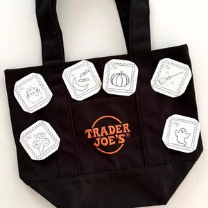 May include: A black canvas tote bag featuring the orange embroidered text Trader Joe's inside a circle. The bag is decorated with six white square patches resembling postage stamps, each displaying black line art of a cauldron, moon, pumpkin, broom, candy, and ghost.