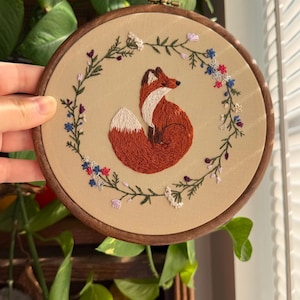 May include: An oval embroidery hoop with a detailed embroidered fox, featuring a fluffy white-tipped tail, surrounded by a floral wreath of blue, red, and purple flowers. The fox is a warm reddish-brown colour, set against a beige fabric.