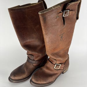 Könnte beinhalten: Ein Paar wadenhohe braune Lederstiefel mit abgenutzter Optik, abgerundeter Zehenpartie und flachem Absatz. Jeder Stiefel verfügt über einen Schnallenriemen am Knöchel und eine verstellbare Schnalle am Schaft sowie ein Browning-Logo an der Seite.