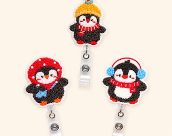 Puede incluir: Tres portatarjetas bordados con forma de pingüino y accesorios de invierno. Un pingüino lleva un gorro amarillo con una flor roja, otro una capucha roja de lunares y el tercero orejeras azules con una bufanda roja. Cada uno tiene un clip.