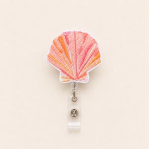 May include: A badge reel featuring an embroidered seashell applique in shades of pink, coral, and orange. The seashell has a white stitched border and is attached to a clear plastic retractable cord housing with a metal clip on the back.