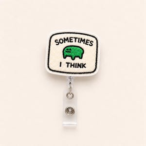 May include: A badge reel featuring a rectangular off-white embroidered patch with black stitched borders. The patch displays the black text "SOMETIMES" above a small green creature illustration and the black text "I THINK" below the creature.