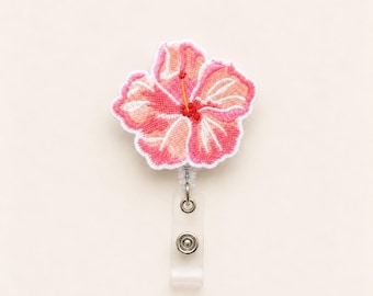 Könnte beinhalten: Ein Ausweishalter mit einem gestickten Hibiskusblüten-Aufnäher in Rosa-, Korallen- und Weißtönen mit gelbem Staubblatt. Die Blüte ist an einem transparenten Kunststoffgehäuse mit einem Metallclip und einem durchsichtigen Vinylriemen befestigt.
