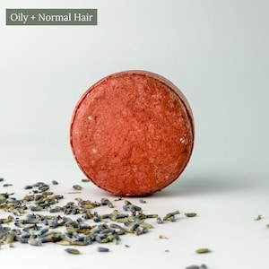 May include: A round, textured, reddish-orange shampoo bar with a label that reads Oily + Normal Hair. The bar is surrounded by scattered dried lavender buds, which are small, elongated, and purple-toned, against a plain light background.