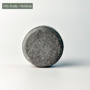 May include: A round, dark gray shampoo bar with a textured surface. The bar is circular and appears solid. Text at the top reads "Oily Scalp + Buildup".