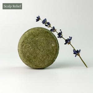 May include: A round, textured olive-green shampoo bar positioned next to a single sprig of dried purple lavender. The text Scalp Relief is visible in the upper left corner of the image. The object has a solid, circular shape with a matte finish.