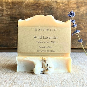 Puede incluir: Una pastilla de jabón Wild Lavender de EDENWILD, de color beige claro y con una etiqueta de papel. La etiqueta dice "Wild Lavender, Tallow + Goat Milk, Sensitive Skin, NET WT 4.5 OZ | 128 G". Una ramita de flores de lavanda en el fondo.