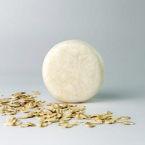 Op de afbeelding: Een rond, solide, gebroken wit shampooblok met een gladde, matte oppervlaktestructuur. Het cirkelvormige blok staat rechtop, omringd door een verspreide verzameling lichtbruine, droge, platte havervlokken op een neutrale ondergrond.