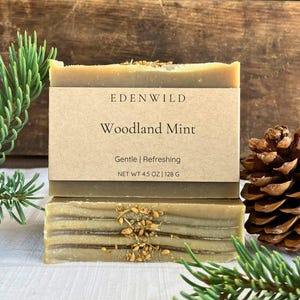 May include: Two bars of Edenwild Woodland Mint soap. The top bar has a brown label with the brand name and product details. The soap is light green with brown layers and topped with dried herbs. A sprig of pine and a pine cone are in the background.