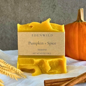 May include: A yellow bar of soap with the text "EDENWILD Pumpkin + Spice Seasonal NET WT 4.5 OZ | 128 G". The soap is next to a small orange pumpkin, dried wheat, and cinnamon sticks. The soap has a textured surface.
