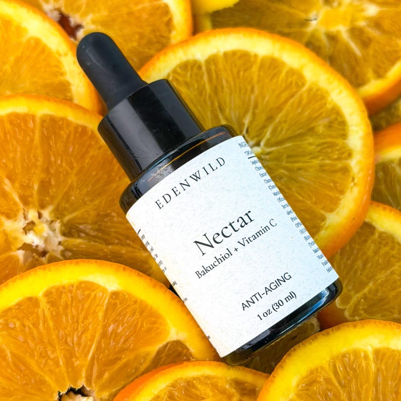 May include: A black glass bottle with a dropper, labeled "EDEN WILD Nectar Bakuchiol + Vitamin C ANTI-AGING 1 oz (30 ml)", rests on a bed of orange slices. The bottle has a white label with black text.