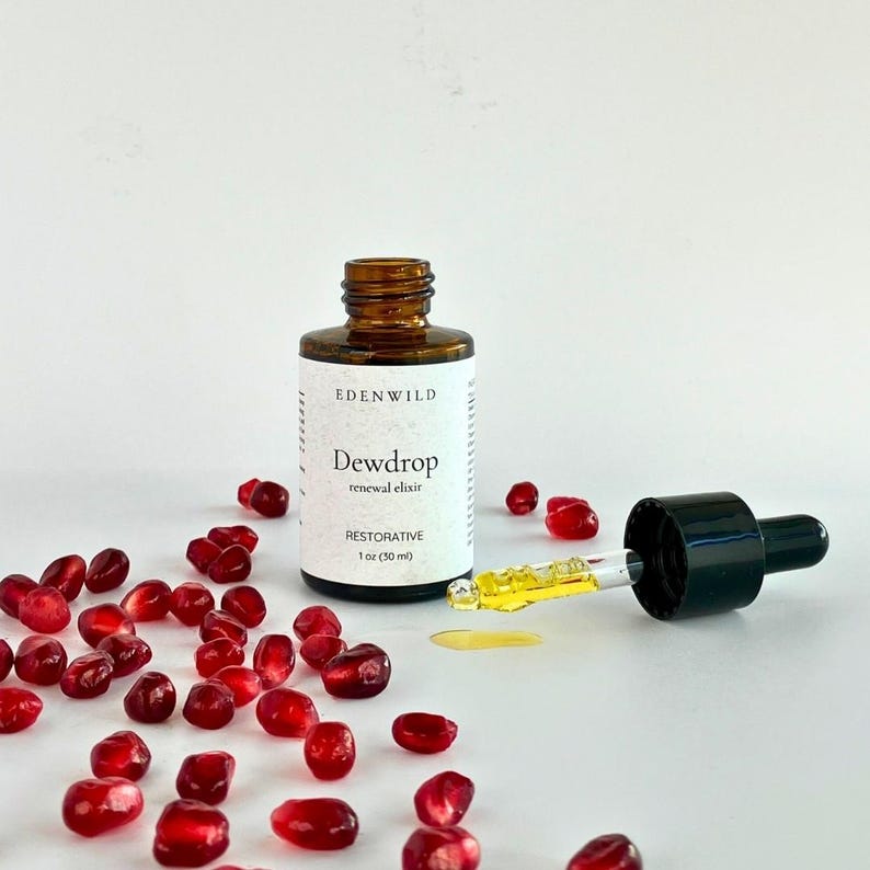 May include: A brown glass bottle of "Dewdrop renewal elixir" with a black dropper and a pool of yellow liquid. The bottle is surrounded by scattered red pomegranate seeds. The label reads "Edenwild" and "Restorative 1 oz (30 ml)".