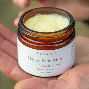May include: A small glass jar of Happy Baby Balm, held in open hands. The jar has a brown tint and a white label with the text "Happy Baby Balm + Colloidal Oatmeal Gentle | Soothe 2 OZ (57 G)". The balm inside is a light yellow colour.