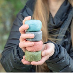 以下が含まれることがあります： 4つのカラフルな丸いシャンプーバーのスタック。一番上のバーは水色、その下にグレー、コーラル、グリーン。ぼやけた緑色の背景の中で、人の手でバーが持たれています。