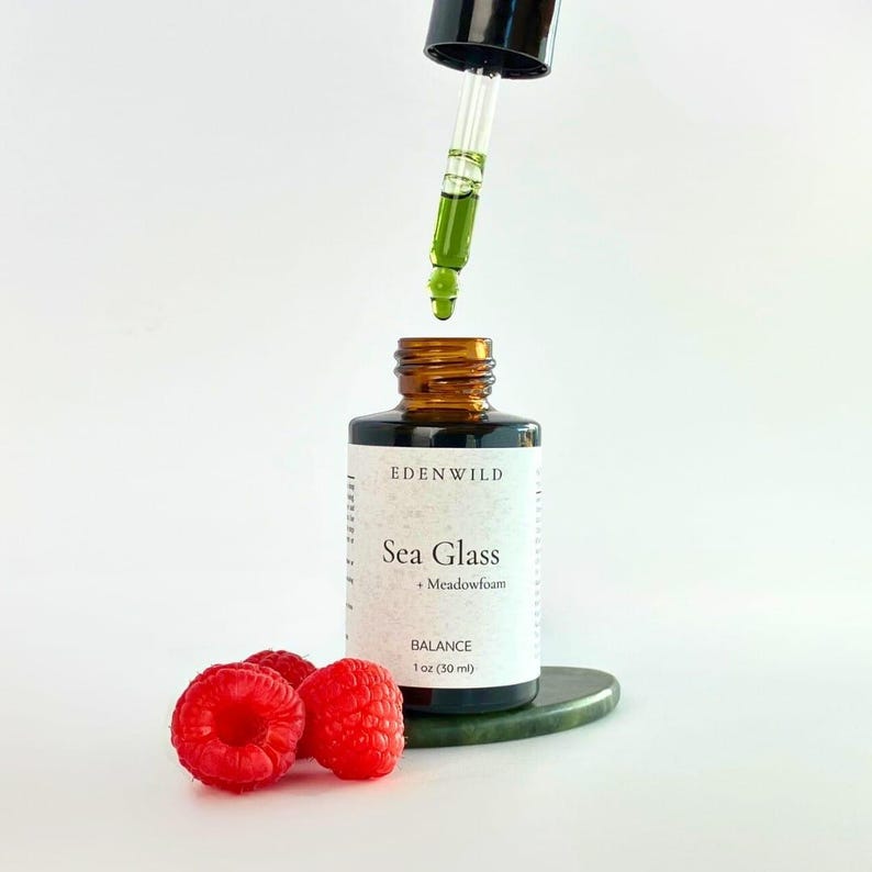 Pu&ograve; includere: Una bottiglia di vetro marrone con un contagocce, etichettata "Sea Glass + Meadowfoam" e "Balance 1 oz (30 ml)." La bottiglia &egrave; accanto a tre lamponi rossi e una pietra verde. Un liquido verde gocciola dal contagocce.