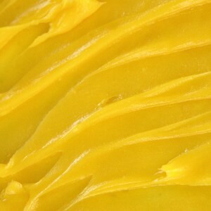 May include: A thick, vibrant yellow substance with a smooth, creamy texture. The surface features distinct, sweeping ridges and peaks created by a tool or utensil, showing a consistent, semi-solid consistency throughout the entire frame.