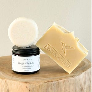 Op de afbeelding: Een rond gebroken wit vast blok, een rechthoekig beige zeepblok met een ingedrukte kolibrie en de tekst EDENWILD, en een kleine donkere glazen pot met een zwarte deksel met het label Happy Baby Balm + Colloidal Oatmeal 2 oz (57 g) van EDENWILD.