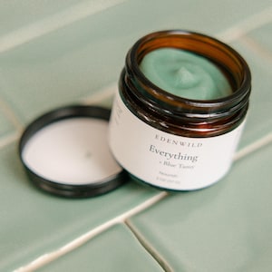 Puede incluir: Un tarro de cristal marrón de crema facial "Everything + Blue Tansy" de Edenwild. El tarro tiene una etiqueta blanca y una tapa negra. La crema del interior es de color verde azulado claro. La tapa está abierta y al lado del tarro sobre una superficie de baldosas verde claro. Contiene 57 g.
