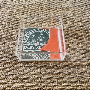 Op de afbeelding: Een heldere acryl dienblad met twee stoffen vierkanten. Eén heeft een grijs en wit bloemenpatroon, de andere is effen oranje. De lade is vierkant.