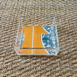 Puede incluir: Una bandeja rectangular de acrílico transparente. El interior de la bandeja presenta un patrón decorativo con formas geométricas naranjas y blancas, y diseños florales azules y blancos. La bandeja se encuentra sobre una superficie tejida y texturizada.