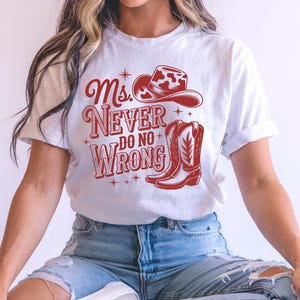 May include: White t-shirt with a red graphic featuring a cowboy hat, boots, and the text "Ms. Never Do No Wrong." The design has a Western theme. The shirt is made of a soft material.