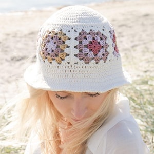 Può includere: Un cappello a secchiello all'uncinetto bianco con accenti floreali a quadretti della nonna nei toni del rosa, viola e beige. Il cappello ha un'ampia tesa e una corona rotonda, ideale per l'estate.