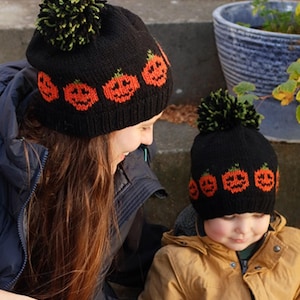 May include: Two black knitted beanies featuring a repeating orange pumpkin pattern around the base and a green and black pom-pom on top. The hats are constructed from a textured yarn and display a consistent design across both sizes shown.