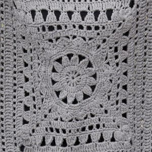May include: Close-up of a gray crocheted square with a floral design in the center. The square features intricate openwork patterns and a textured surface. The crochet work is detailed and the color is a solid gray.