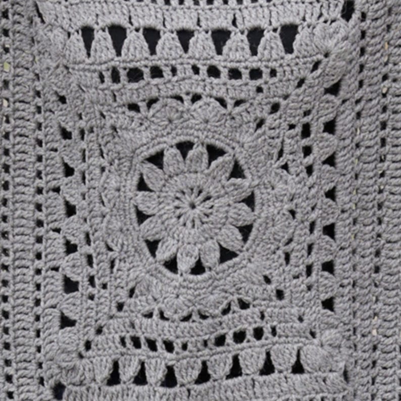 May include: Close-up of a gray crocheted square with a floral design in the center. The square features intricate openwork patterns and a textured surface. The crochet work is detailed and the color is a solid gray.