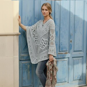 Op de afbeelding: Een lichtgrijze, open geweven gehaakte poncho met een V-hals en klokmouwen. De poncho heeft ingewikkelde bloemen- en geometrische patronen. De persoon draagt een grijze spijkerbroek en houdt een sjaal met patroon vast. De achtergrond is een blauwe deur.