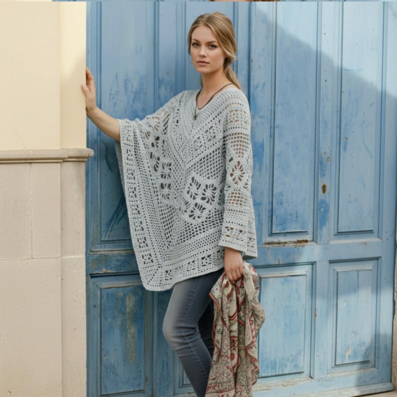 Op de afbeelding: Een lichtgrijze, open geweven gehaakte poncho met een V-hals en klokmouwen. De poncho heeft ingewikkelde bloemen- en geometrische patronen. De persoon draagt een grijze spijkerbroek en houdt een sjaal met patroon vast. De achtergrond is een blauwe deur.